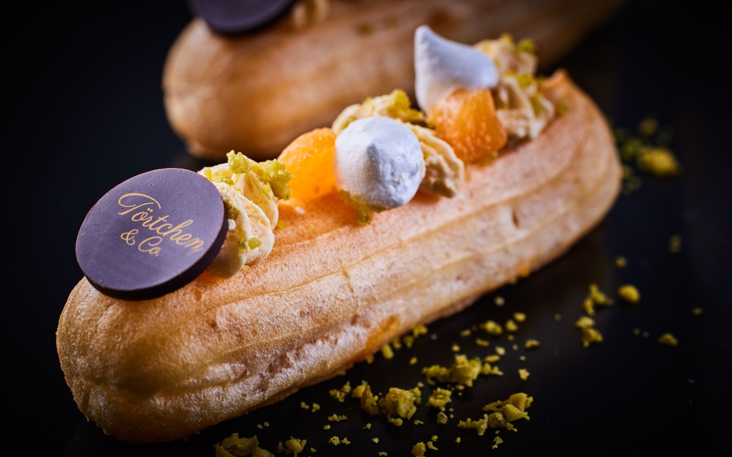 Eclair mit Creme, kandierten Zitrusfrüchten, Pistazienstückchen und einer Schokoladenplakette mit dem Schriftzug ‚Törtchen & Co‘.